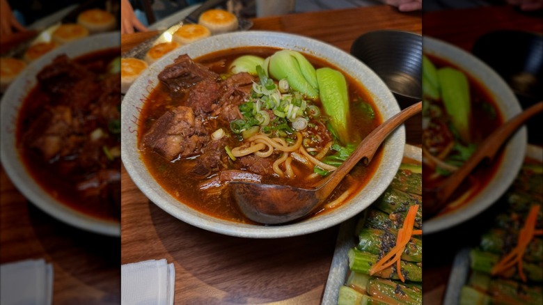 A large bowl of soup with dark broth, noodles, meat, and greens placed on a wooden table.