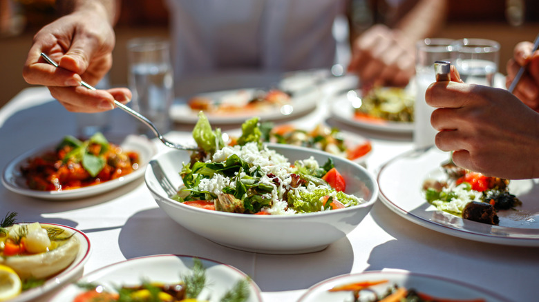 Two people eating Greek-style salads and appetizers on white cloth table in a sunny restaurant.