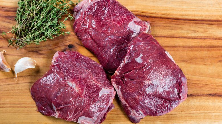 Raw beef cheeks on wooden cutting board alongside sprigs of thyme and cloves of garlic