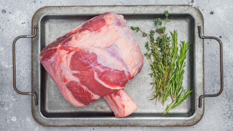 Raw whole beef shank on metal pan alongside sprigs of thyme and rosemary
