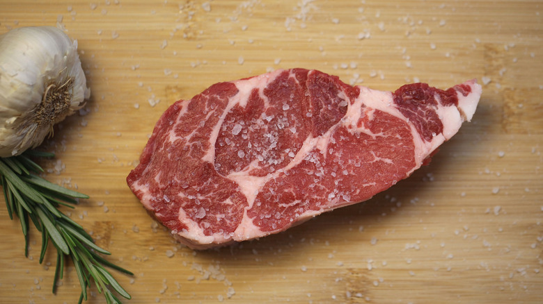 Raw chuck eye steak sprinkled in salt alongside sprig of rosemary and bulb of garlic