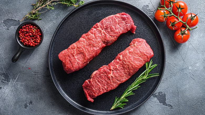 Two raw Denver steaks on black plate alongside a sprig of rosemary
