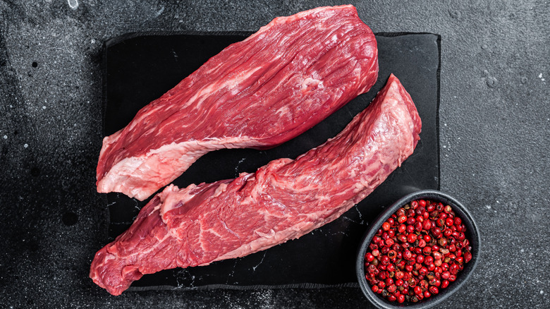 Two raw bavette flap steaks on black stone board alongside a bowl of pink peppercorns