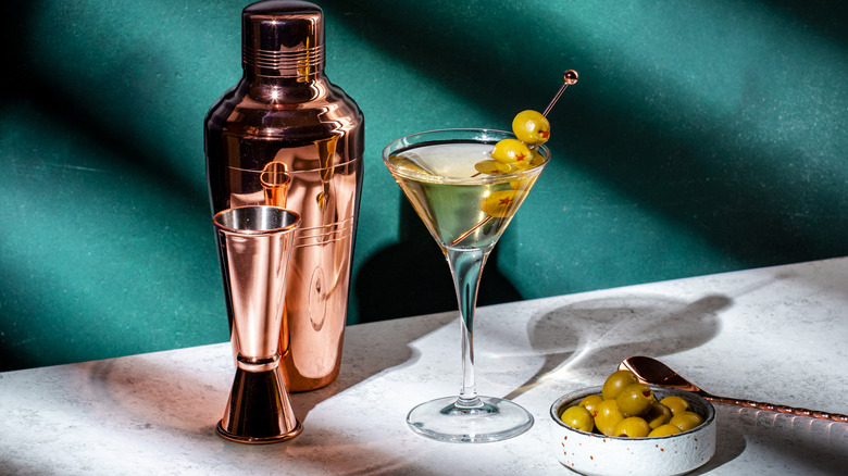 A vodka martini with olives on a table next to a cocktail shaker, a jigger, and a tin of olives.