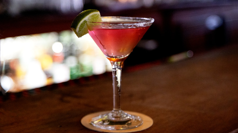A Cosmopolitan in a martini glass, garnished with a lime wedge, sitting on a coaster on a bar.