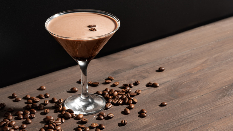 An Espresso martini on a wooden table surrounded by coffee beans.