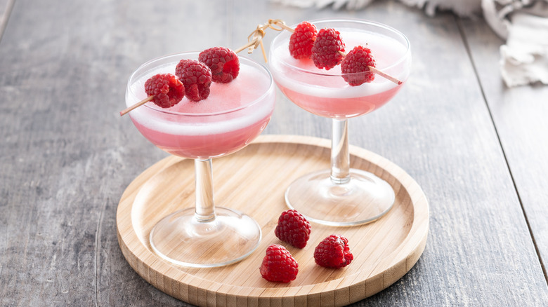 Two French martinis in coupe glasses, garnished with raspberries on a wooden tray with three raspberries.