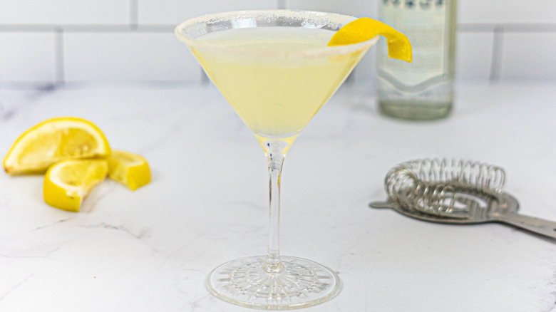 A Lemon drop on a counter next to a strainer and lemon wedges.