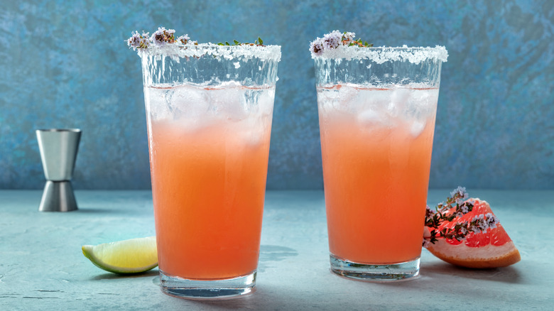 Two salty dogs side by side next to a grapefruit wedge, lime wedge, with a jigger in the background.