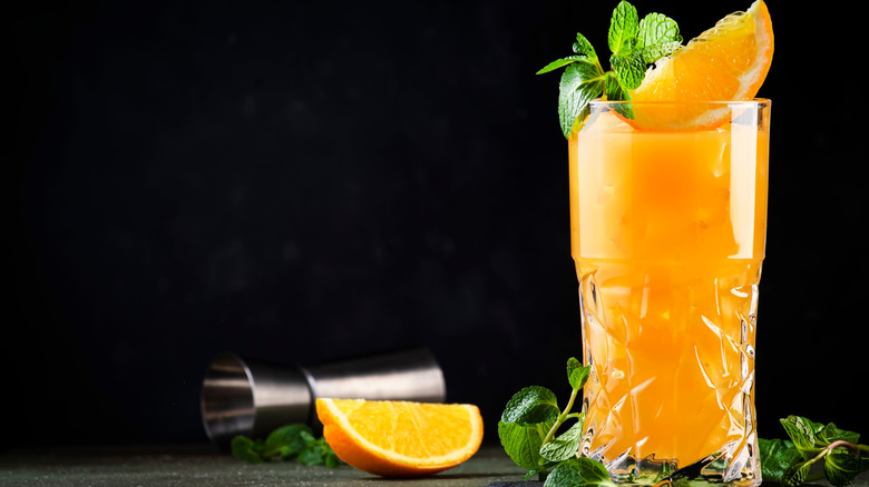 A screwdriver cocktail in a tall glass, garnished with an orange wedge and mint leaves next to another orange wedge and a jigger.
