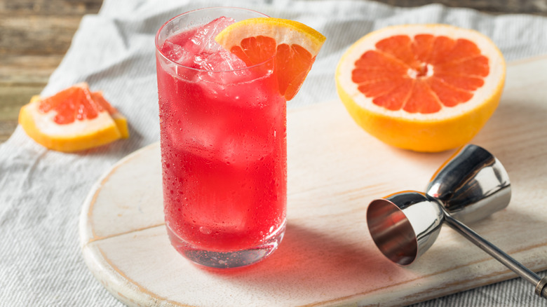 A sea breeze garnished with a grapefruit wedge on a wooden board with a grapefruit half and wedge and a jigger.