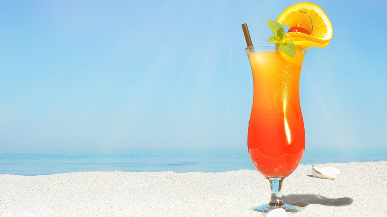 A Sex on the beach cocktail garnished with an orange slice and a cherry sitting on a sandy beach.