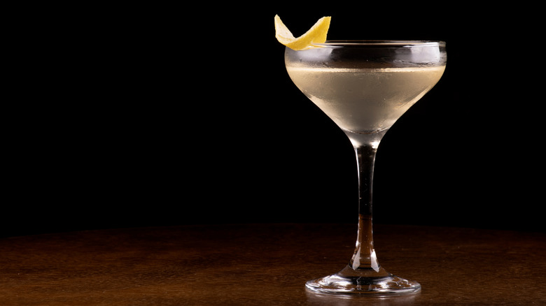 A Vesper cocktail in coupe glass with lemon garnish on a table against a dark background.