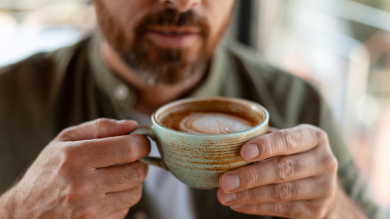 A man holds an artfully crafted mug filled with coffee in front of him ready to drink.