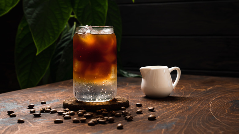 An iced espresso soda drink next to a small milk jug on a wooden table.