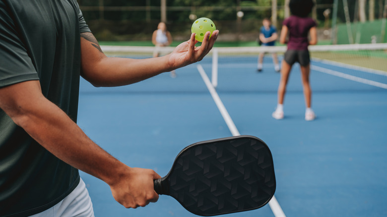 Man ready to serve with a pickleball racquet in one hand and the ball in the other, with three other players on the court