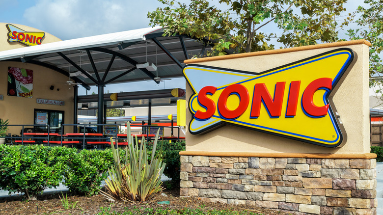 Sonic sign on a brick base next to small bushes with the fast food restaurant in the background