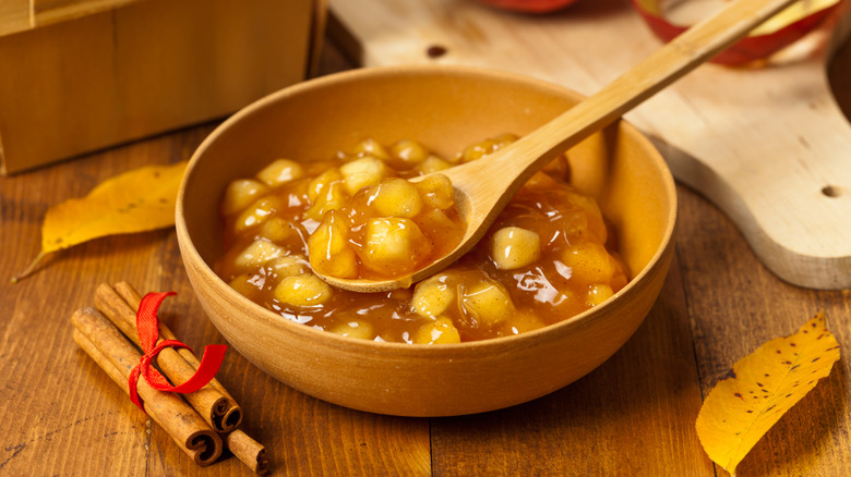 A bowl of apple pie filling next to a bundle of cinnamon sticks and scattered leaves.