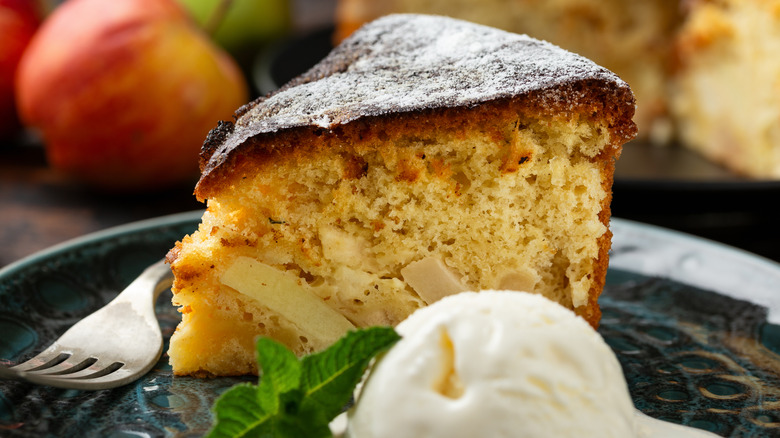 A slice of apple cake dusted with powdered sugar on a plate with a scoop of vanilla ice cream and a sprig of mint.