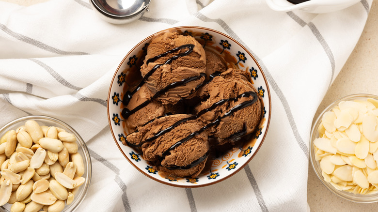 Bowl of chocolate ice cream and chocolate syrup alonside bowls of peanuts and almonds