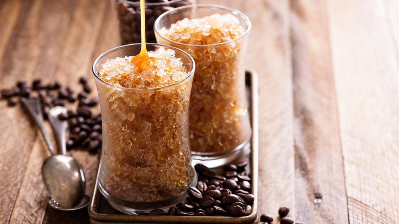 Two glasses of coffee granita on a small rectangular metal tray on a wooden surface with scattered coffee beans and two spoons