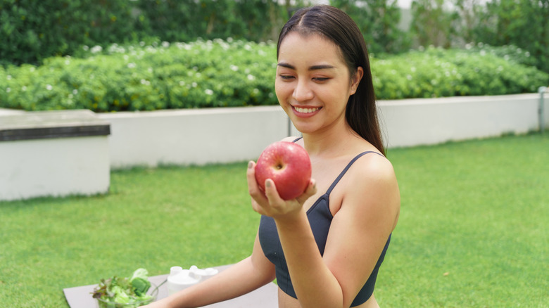 A woman sitting on a yoga mat in the grass holding a red apple