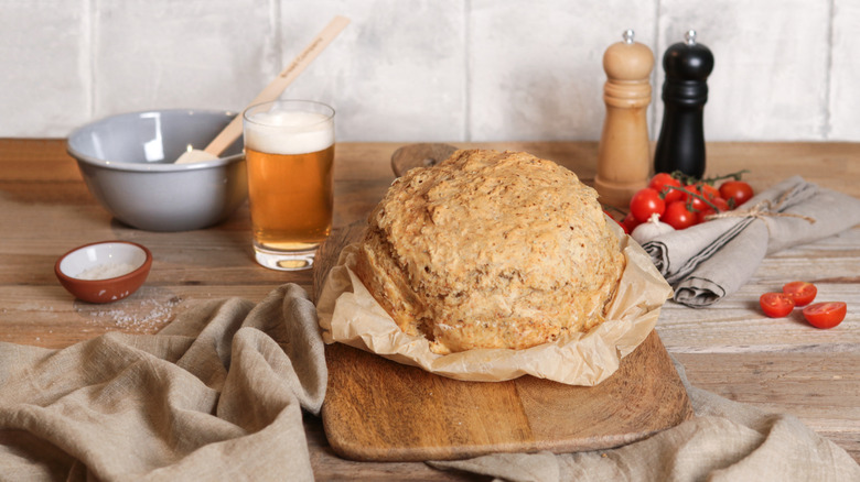 A loaf of beer bread next to a glass of beer, tomatoes, a bowl, and seasonings.