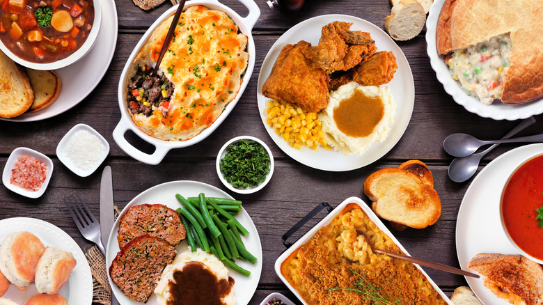 A medley of comfort food dishes displayed on a wooden table including chicken pot pie, meatloaf, fried chicken, stew, shepherd's pie, and mac and cheese.