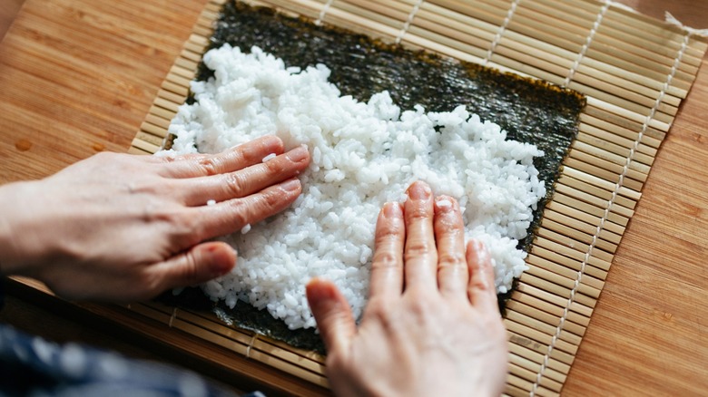 Person putting rice on nori for sushi, all of which sits on a bamboo sushi rolling mat.