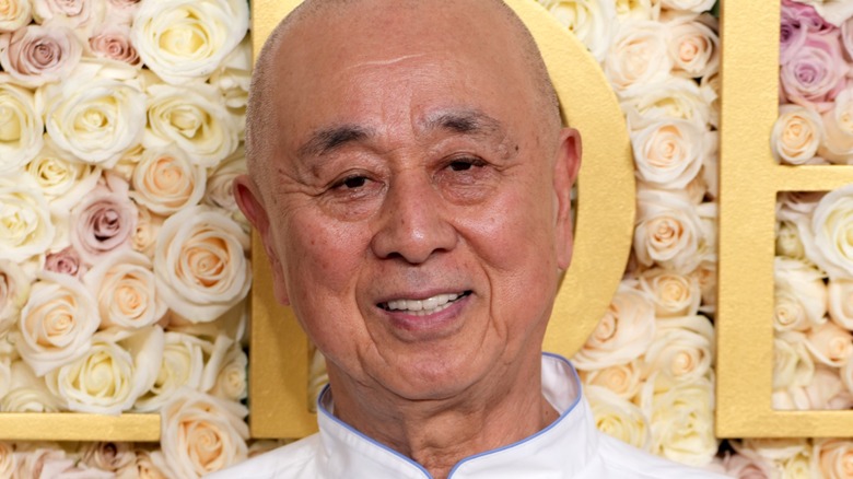 Chef Nobu Matsuhisa posing for photograph in front of a wall of white and pink roses.