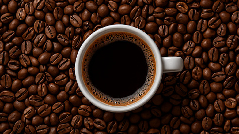 A white cup of coffee in the middle of a pile of coffee beans that surround it.