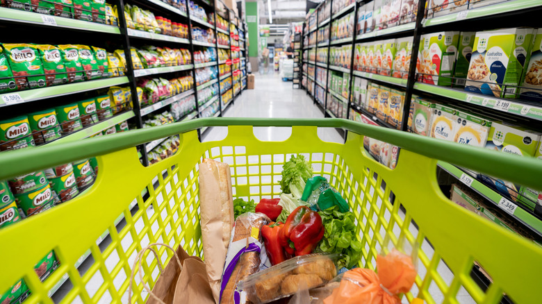 Green plastic shopping cart full of produce and bread in grocery aisle