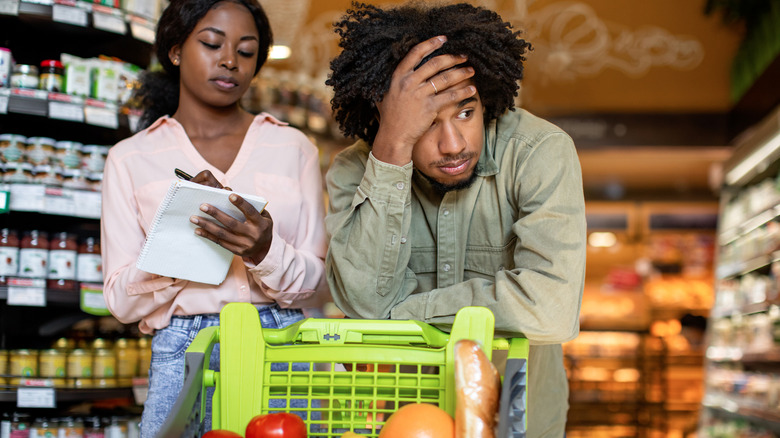 Frustrated man leaning on grocery cart and a woman marking off grocery list