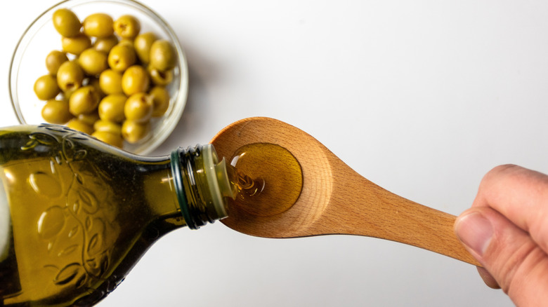 A bottle of extra virgin olive oil is poured into a wooden spoon above a bowl of olives