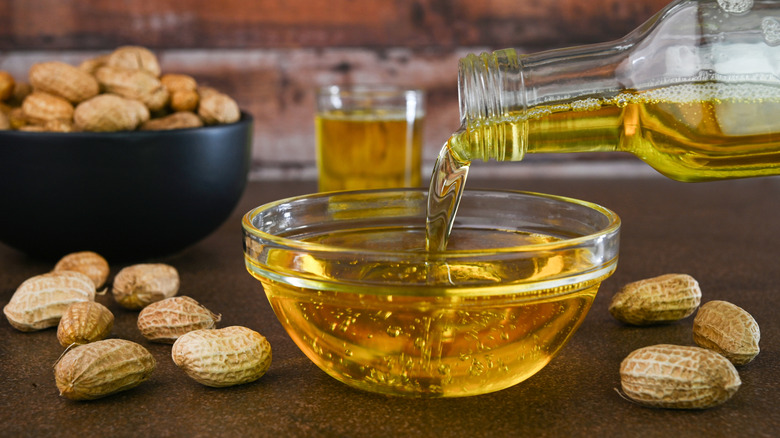 A glass bottle of peanut oil pours oil into a glass dish, surrounded by peanuts.