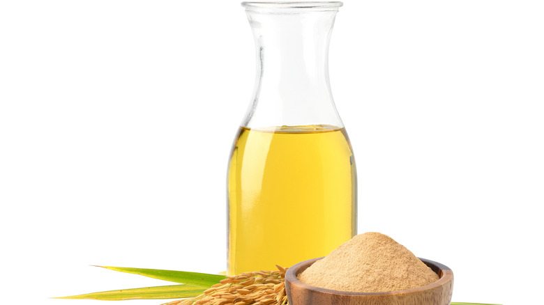 A glass bottle of rice bran oil on a white background with a wooden bowl of bran flour