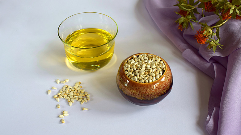 A glass bowl of safflower oil and a ceramic bowl of safflower seeds on a white table, flowers on a cloth to the side