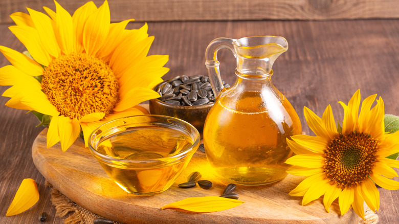 Sunflower oil sits on a wooden cutting board, surrounded by sunflowers and a bowl of sunflower seeds.