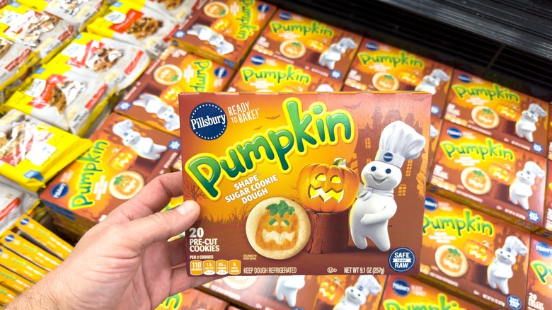 A hand holds up a box of Pillsbury Ready to Bake Pumpkin Shape Sugar Cookie Dough above a refrigerated case of more boxes
