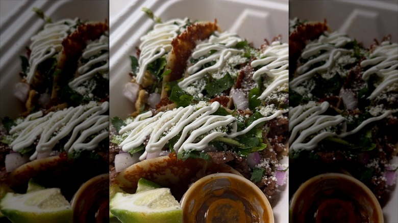 Three crispy tacos topped with sour cream, cilantro, and fresh onions in a to-go container