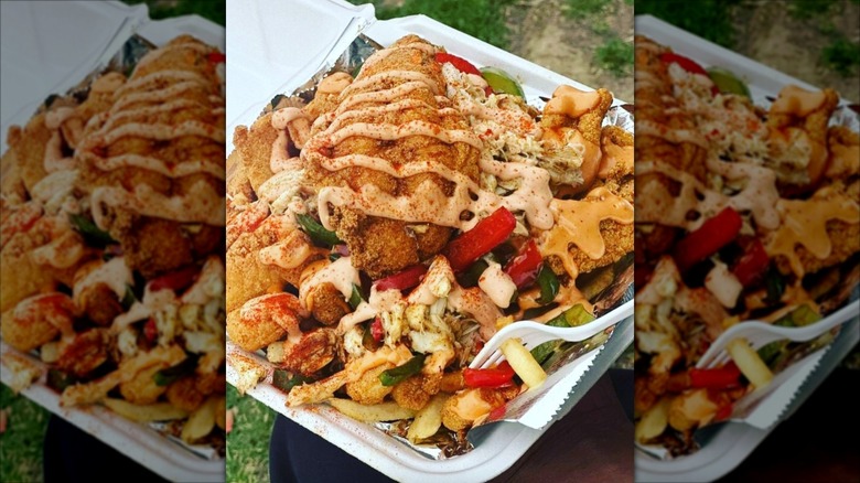 Seafood, onions, and peppers loaded onto a bed of fries topped with a deep fried lobster tail and seafood sauce in a to-go container lobster tail and christened with secret Gawds seafood sauce