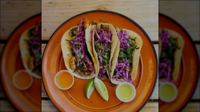 Three shrimp tacos with shredded red cabbage, cilantro, two dipping sauces, and lime wedges on an orange plate