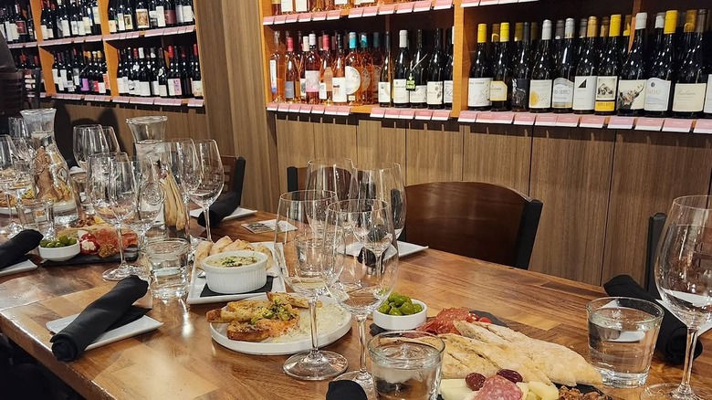Long wooden table with appetizers and wine glasses and shelves of wine in the background at Corkscrew Wine and Cheese
