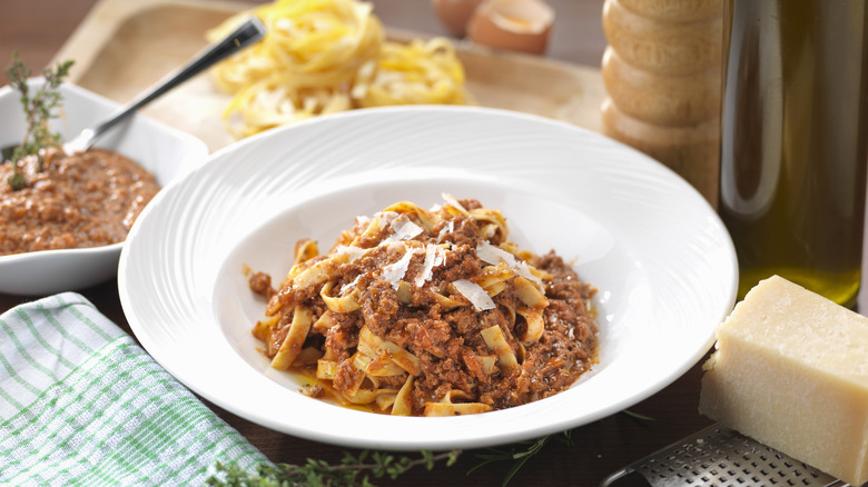 Bowl of pasta bolognese in a white bowl, surrounded by ingredients, seasonings, and garnishes.