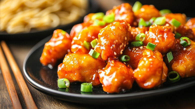 Shiny breaded chicken nuggets in sweet and sour sauce with sliced scallions sprinkled on top on a black round plate