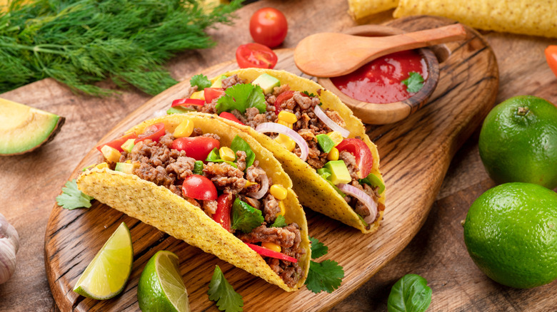 Tacos with a crunchy corn shell on a wood board next to limes, tomatoes, and fresh herbs