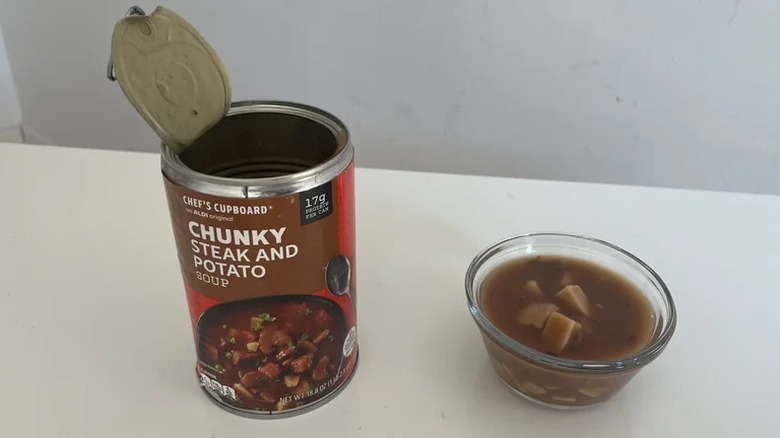 An open can of Chef's Cupbaord Chunky Steak and Potato soup next to a small ramekin of the soup on a table.