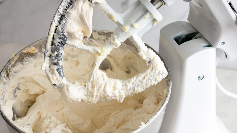 A stand mixer covered in buttercream above a metal bowl full of mixed buttercream.