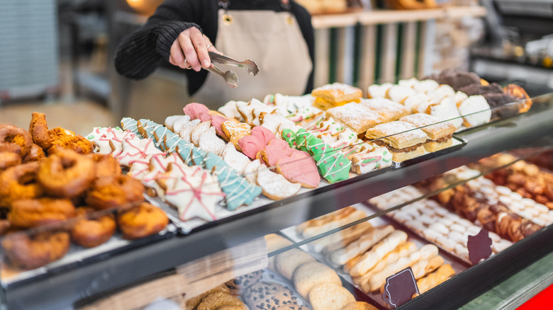 A worker reaches for baked goods with tongs behind a case filled with a variety of pastries and cookies