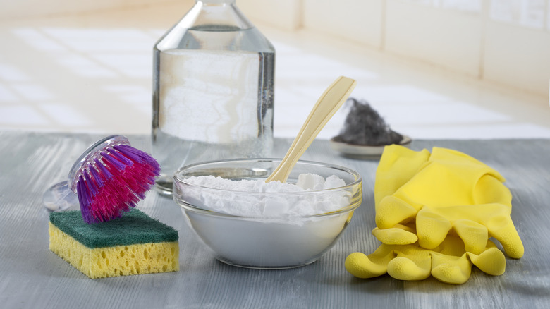 A glass bowl of baking soda next to a sponge and rubber gloves, with a bottle of vinegar in the background.
